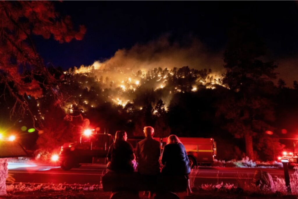 A family watches a raging wildfire across the mountain at night with fire trucks nearby — wildfire home protection matters