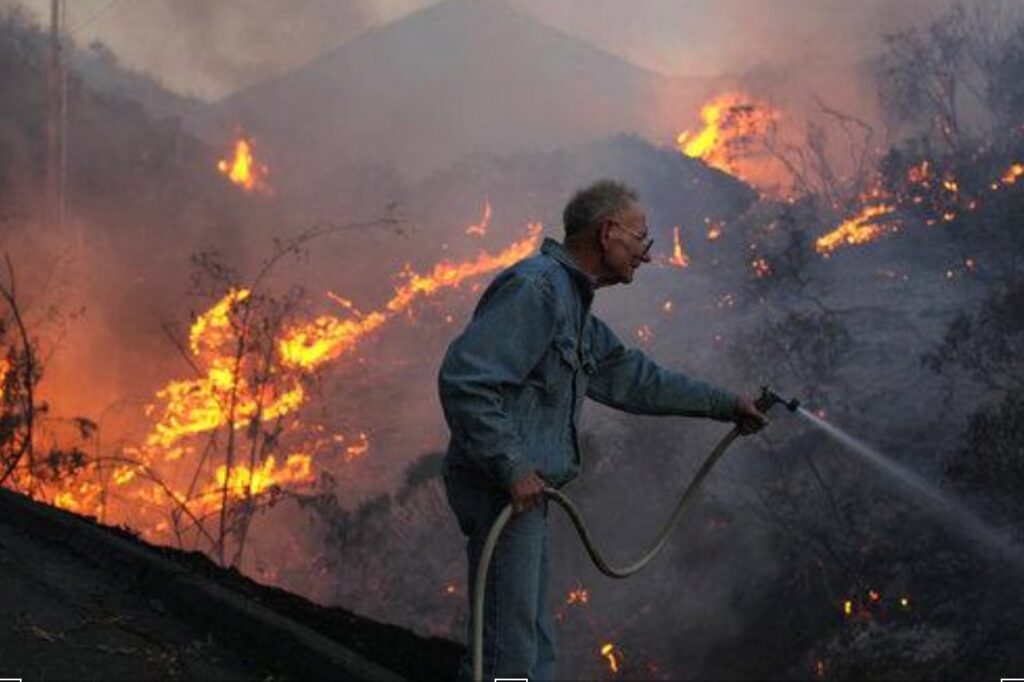 Man using hose as wildfire burns through dry hillside